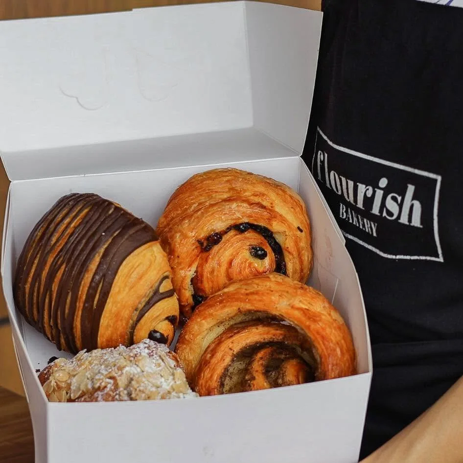 A white box of croissants and scrolls held beside a Flourish Bakery apron